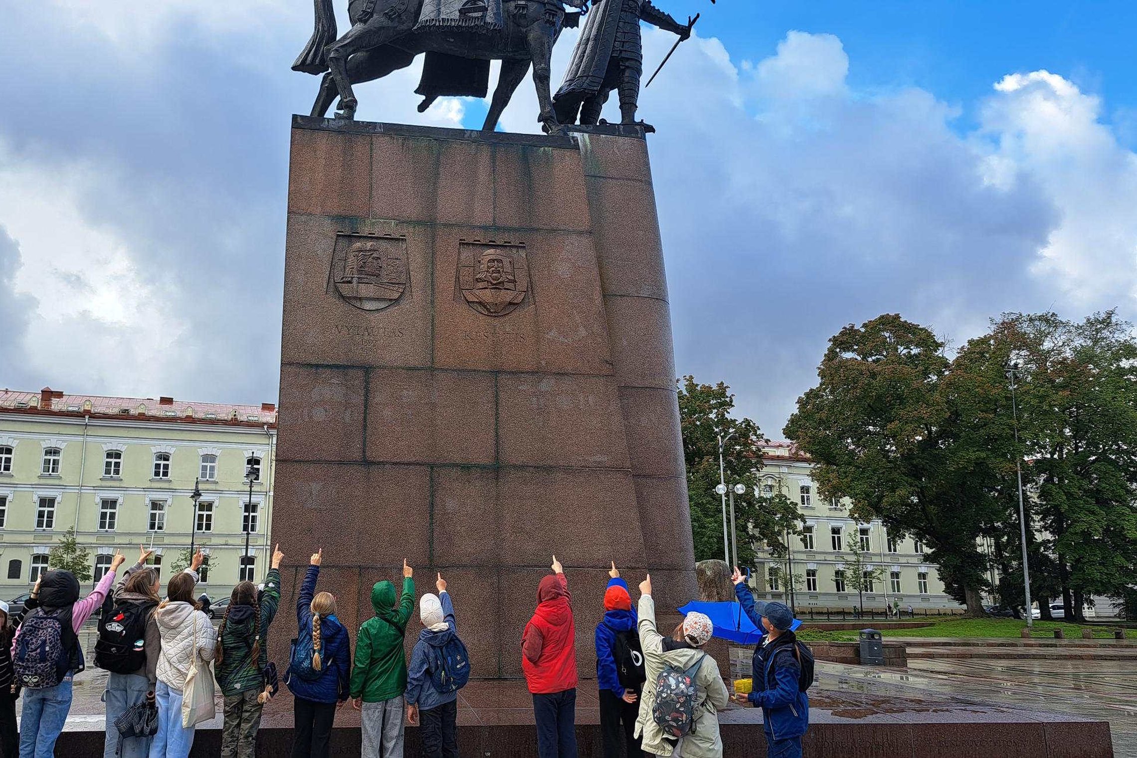 Grupė penktokų iš Sietuvos gimnazijos stovi prie Gedimino paminklo Vilniuje ir rodo rankomis į viršų, link skulptūros.