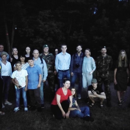 Nemažas ekskursijos dalyvių būrys stovi naktyje ir pozuoja nuotraukoje. Aplinkui naktis, labai tamsu. Žmonės vasarariškai apsirengę, grupėje yra ir vaikų. Taip pat yra ir kariškių su karinėmis uniformomis.