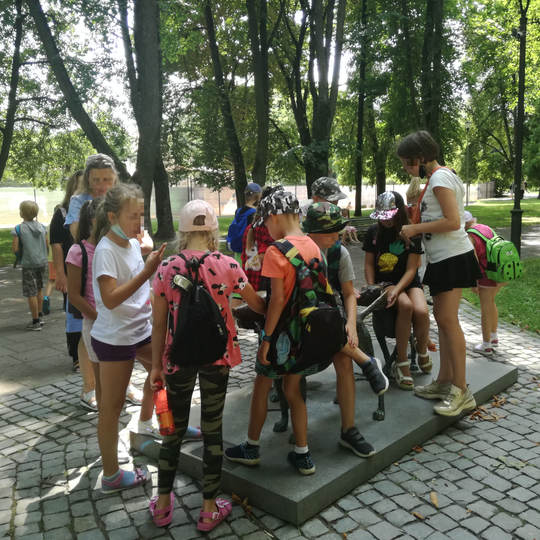Ekskursijos dalyviai šalia lietuvių skalikų skulptūros. Skulptūros yra Pilies parke. Vaikai apsirengę vasariškais rūbais, yra karšta diena.