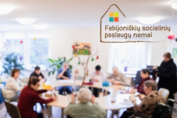 Nuotraukoje prie apvalaus stalo sėdi žmonės, kurie sėdi ant kėdžių arba neįgaliųjų vėžimėliuose. Vyksta edukacinė veikla.