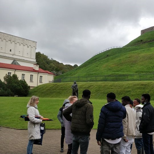 Būrelis tamsiaodžių vaikinų apsupę šviesiaplaukę moterį klausosi. Toliau įspūdingai atrodo žalia veja apaugęs, kaskadomis besileidžiantis Gedimino kalno šlaitas, jo papėdėje paminklas Vytautui Didžiajam. Ant Gedimino kalno matyti keturkampiai Aukštutinės