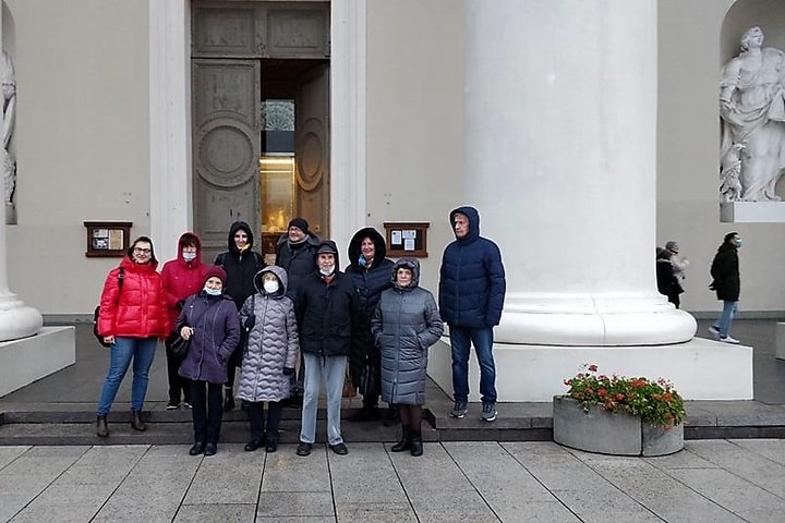 Ekskursijos dalyviai stovi ties Vilniaus katedros centrinio įėjimo tarp kalonų. Dalis žmonių yra su veido kaukėmis. Yra vėsi diena, žmonės šiltai apsirengę.