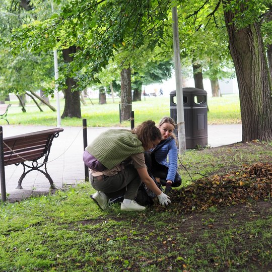Dvi moterys parke renka lapus į krūvą šalia suoliuko.