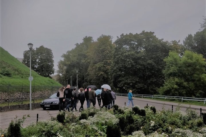 Lietinga diena, pilkas apsiniaukęs dangus. Žmonių grupė dalyvauja ekskursijoje. Kai kurie eina su su lietsargiais. Žmonės su gidu eina pro nuostabiai žydintį gėlyną. Tai Gedimino kalno papėdė. Toliau auga medžiai. Jie dar su žaliais lapais, nors tai rugs