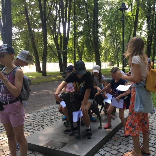 Vaikai apgulę lietuvių skalikų skulptūrą Pilių parke. Vaikai rankose turi popieriaus lapus, daugelis dėvi kepures nuo sauės.