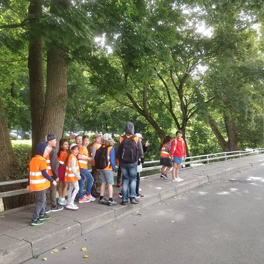 Nuotraukoje matome šaligatviu einančių dvylikos vaikų grupę su vienu lydinčiu asmeniu. Jie visi yra apsirengę ryškiaspalves šviesą artspindinčias liemenes. Pats vyriausias asmuo nuotraukoje nešasis juodą kuprinę. Vienas vaikas yra pasislėpęs už vyriausio