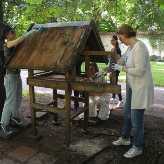 Žmonės parke valo ir tvarko medinę lesyklą po medžiais.