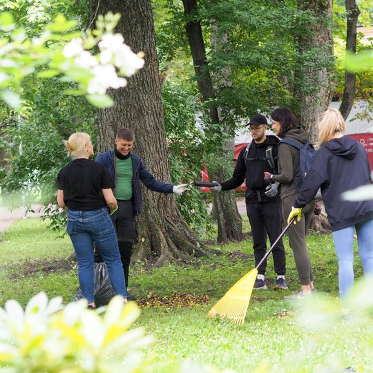 Žmonės parke dalyvauja talkoje, grėbia lapus ir tvarko aplinką.