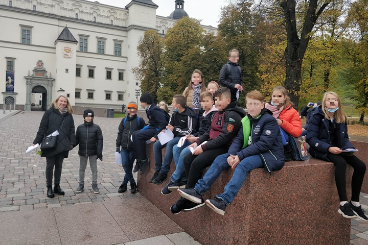 Vaikų grupė ilsisi susėdę ant aukštų granitinių kubų. Šalia stovi mokytoja su vienu vaiku ir laiko jo ranką. Už jų yra senas parkas ir Valdovų rūmai.