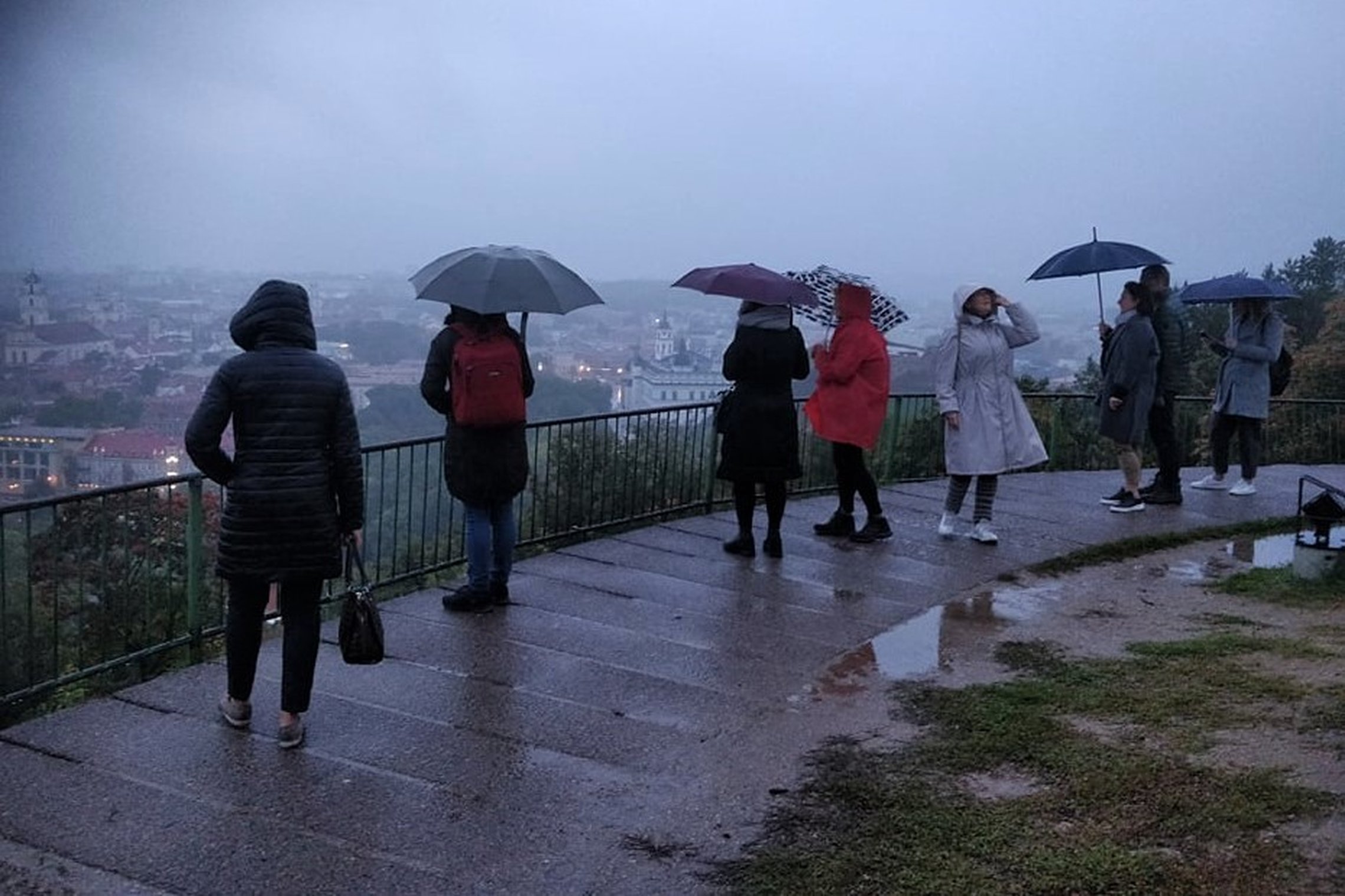 Lietingas oras, žmonės su lietsargiais stovi prie Trijų Kryžių paminklo ir žiūri į miestą. Miestas paskendęs lietuje ir vakaro prietemoje.