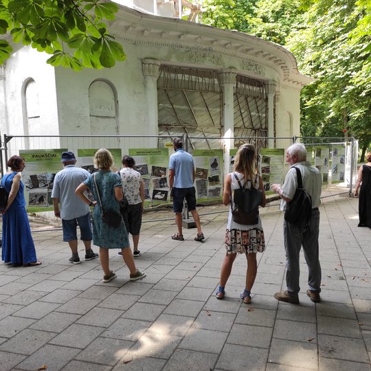 Pilių parke vyksta Vilniaus pilių paukščių nuotraukų parodos atidarymas. Susirinkę būrelis žmonių. Yra išdėliotos kėdės ir stalas. Aplinkui seni medžiai.