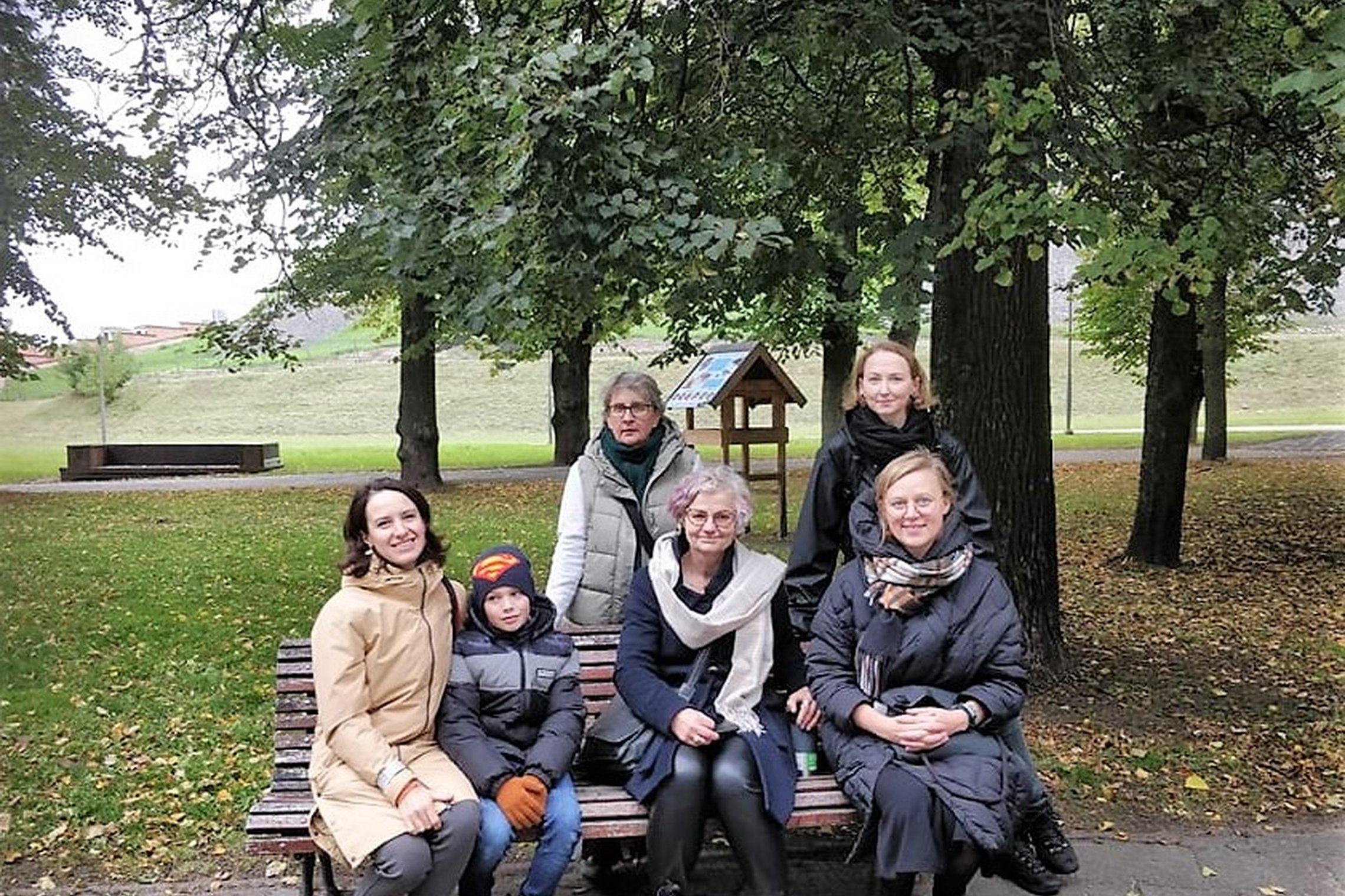 Nedidelė grupelė žmonių sėdi Pilių parke ant suolo ir pozuoja nuotraukai. Tai ekskursijos alyviai.