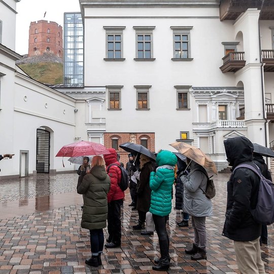 Ekskursija su gide yra Valdovų rūmų kieme. Nuotraukos kairėje matosi Gedimino pilies bokštas. Gidė kažką rodo ranka, žmonės įdėmiai klausosi. Vis dar lyja, todė žmonės yra su skėčiais.