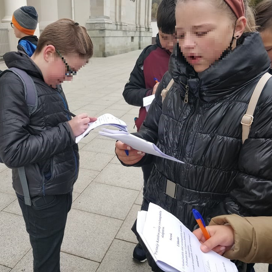 Vaikai turi rankose užduočių lapus. Tai orietacinis žaidimas, jie žaidžia. Berniukas nuotraukos centre rašo į užduotis, o mergaitė, stovinti šalia, garsiai skaito.