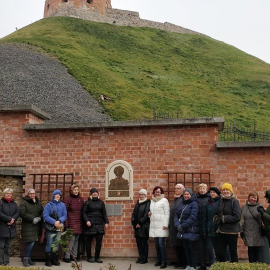 Nemaža žmonių grupė pozuoja prie sienos iš plytų. Ant sienos kabo Bonos Sforzos bareljefas. Visa tai yra Gedimino kalno papėdėje.