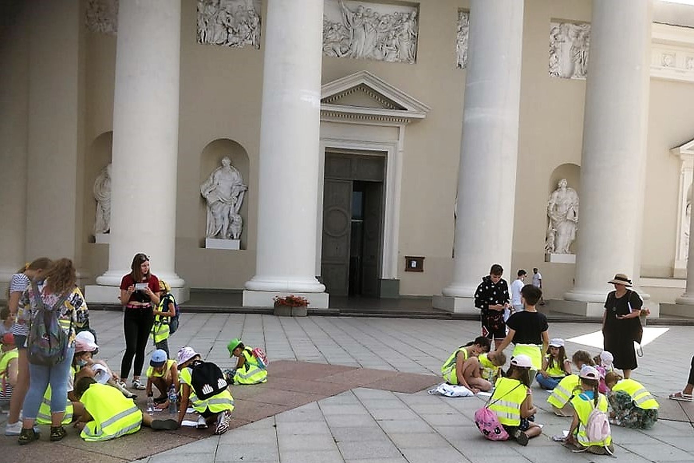 Ryškiomis, šviesą atspindinčiomis liemenėmis dėvintys vaikai žaidžia orientacinį žaidimą Vilniaus pilių rezervate priešais katedros centrinį įėjimą. Jie sutūpę ant šaligatvio sprendžia užduotis.
