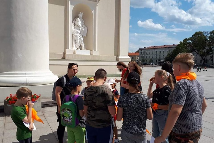 Vaikų grupė prie Vilniaus katedros centrinio fasado. Jiems yra pasakojama pilių istorija ir vaikai klausosi.