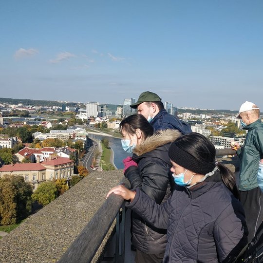 Ekskursijos dalyviai Gedimino pilies bokšto terasoje. Daug giedro dangaus, nei debesėlio. Jie žiūri į apačią. Gražus Vilniaus miesto vaizdas tesiasi į tolį.