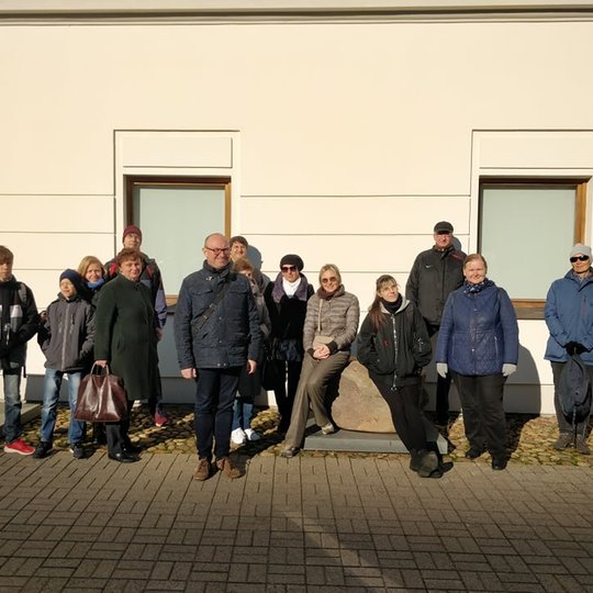 Nemaža ekskursijos dalyvių grupė paozuoja nuotraukoje prie pastato sienos.