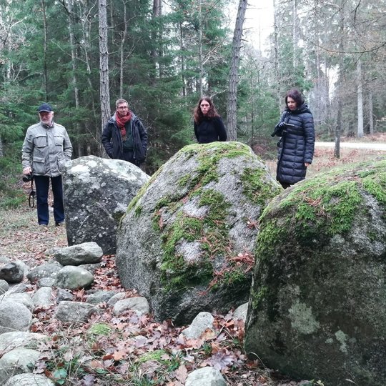 Penki žmonės stovi miške prie didelių, samanomis apaugusių riedulių. Aplink juos išsibarsčiusios mažesnės akmenų nuolaužos, žemę dengia rudeniški lapai. Fone matyti spygliuočių medžiai ir siauras keliukas.