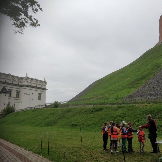 Būrelis vaikų šaudo iš lankų Gedimino kalno papėdėje. Dešinėje amt kalno stovi Gedimino pilies bokštas. Vaikai dėvi ryškias ir vienodas padidinto matomumo liemenes.