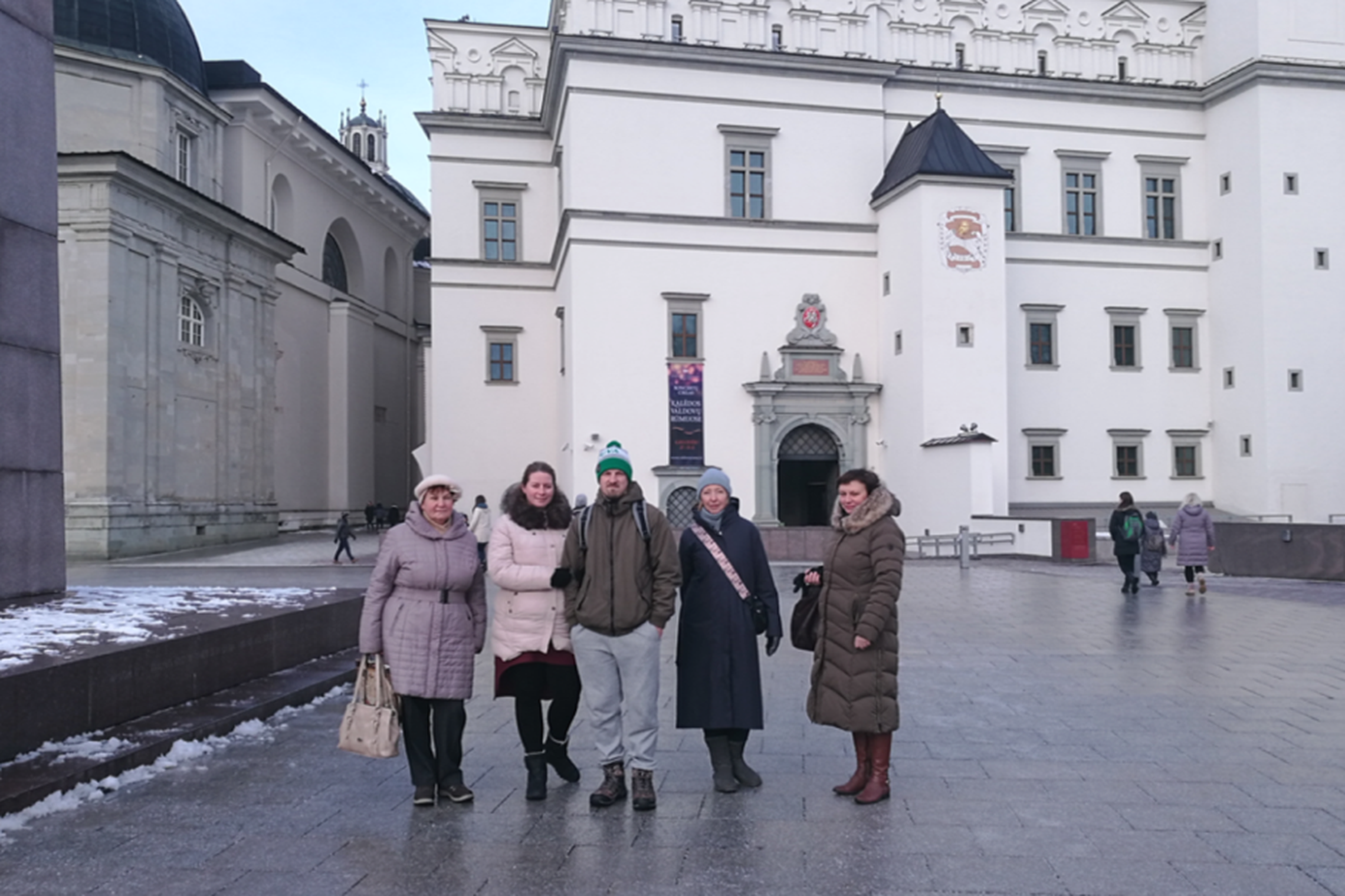 Penkių žmonių grupė stovi netoli Gedimino paminklo ir Vadovų rūmų. Žmonės šiltai apsirengę. Nuotraukos kairėje yra Vilniaus katedra. Žiemos laikas, matosi sniegas.