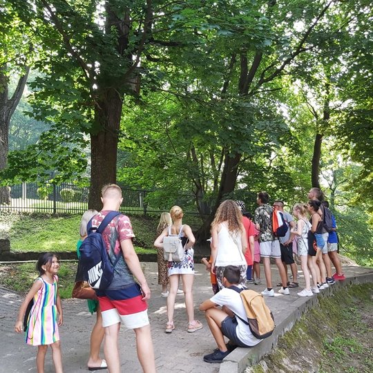 Vaikai dalyvauja ekskursijoje. Uksmingame parke, kur seni medžiai ir apvėsis, vaikai klauso ką pasakoja gidė.
