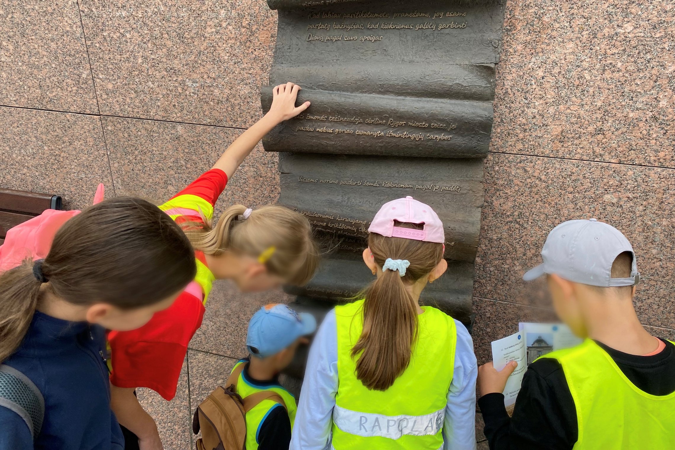 Vaikai su geltonomis liemenėmis tyrinėja Gedimino laiško skulptūrą Vilniuje, skaito užrašus ir ieško atsakymų edukacijos metu