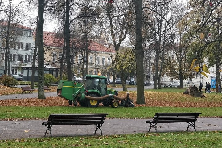 Senas parkas, kurio vejomis važiuoja nedidelis traktorius. Jis su specialiu įrenginiu siurbia rudeninius lapus. Tai priešpaskutiniai lapai, nes ant medžių dar kabo šiek tiek lapų. Nuotraukos centre yra parko suoliukai šalia parko tako. Už parko matosi se