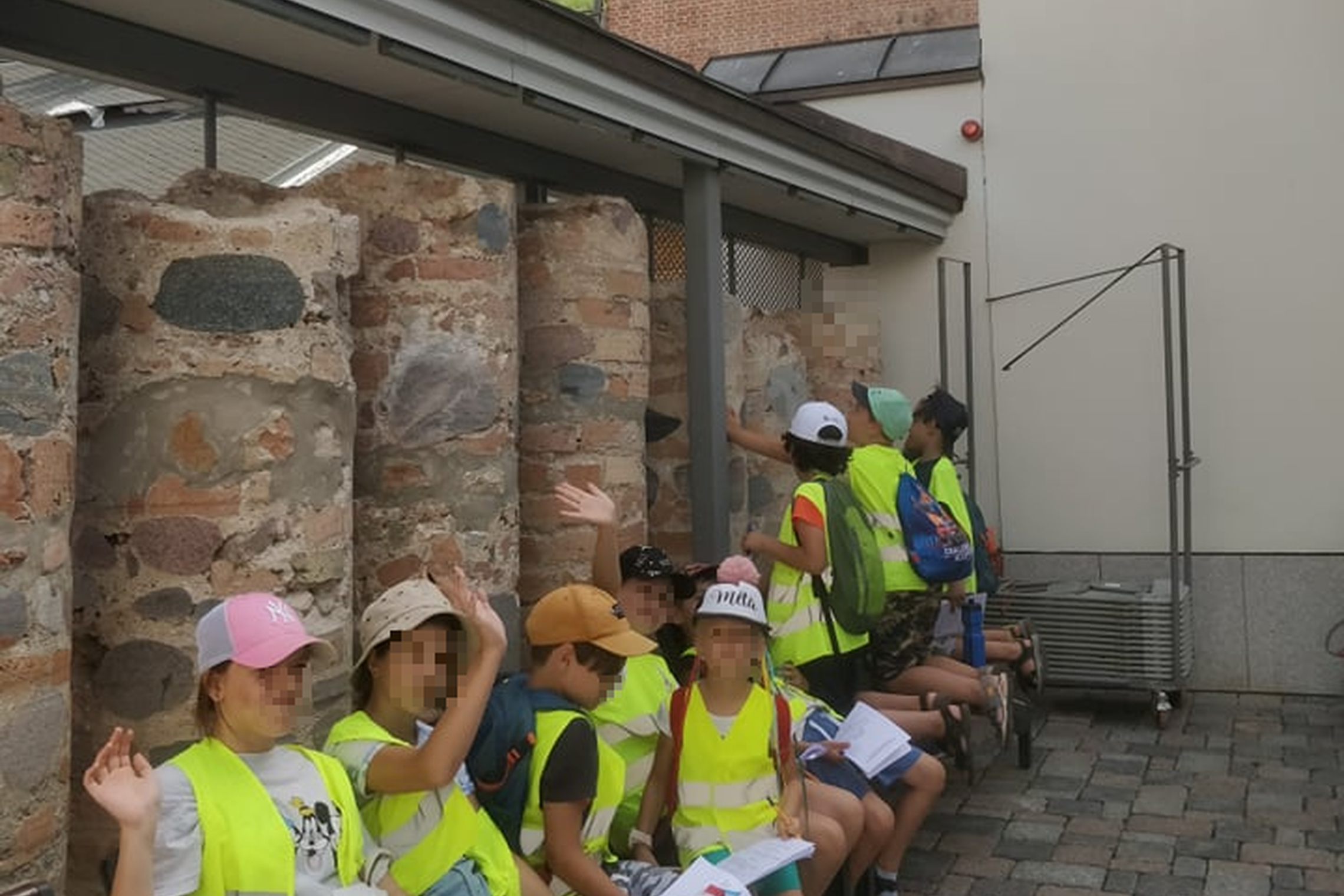 Stovyklos vaikai sėdi Valdovų rūmų kieme šalia senovinių mūro kernų. Nuotraukos centre matosi Gedimino pilies kalnas ir Gedimino bokštas.