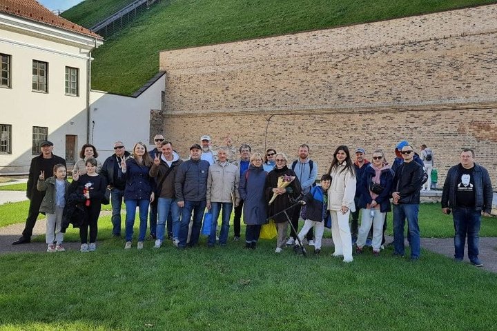 Didelė ekskursijos grupė pozuoja prie šiaurinės Gedimino kalno papėdės. Už jų yra kalno šlaitą laikanti išmūryta siena.