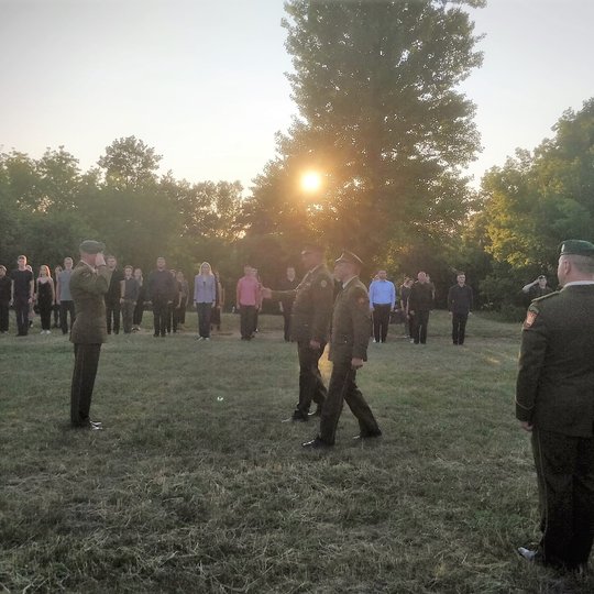 Priešais šaulių naujokus, kurie išrikiuoti į vieną eilę, vyksta karininkų pasisveikinimas. Trys šaulių vadai vienas kitam atiduoda pagarbą. Vakarinė saulė šviečia pro medžių šakas.