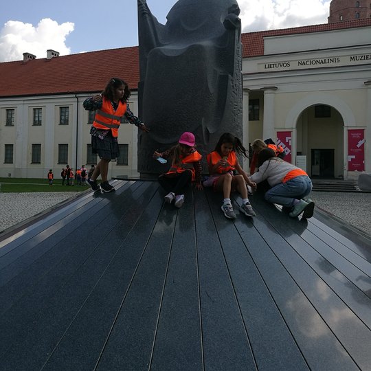 Vaikai žaidžia aplink Mindaugo paminklą. Mindaugo paminklas yra iš tamsios spalvos granito. Vaikai dėvi šviesą atspindinčias apsauginęs liemenes.