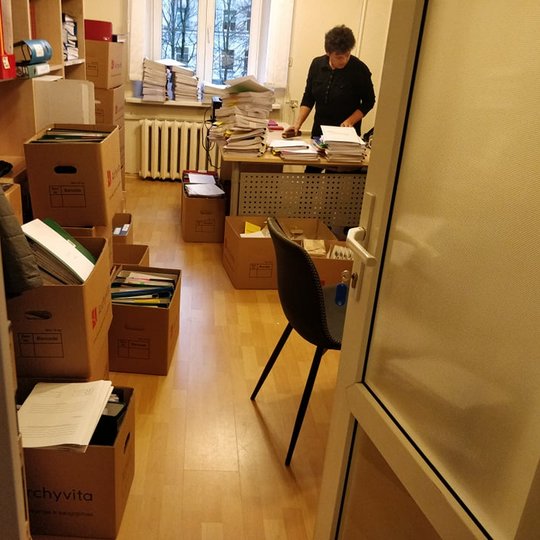 Nuotraukoje kabinetas, kuriame dirba moteris. Ji rušiuoja dokumentus, dėlioja segtuvus. Dokumentai ruošiami archyvuoti.