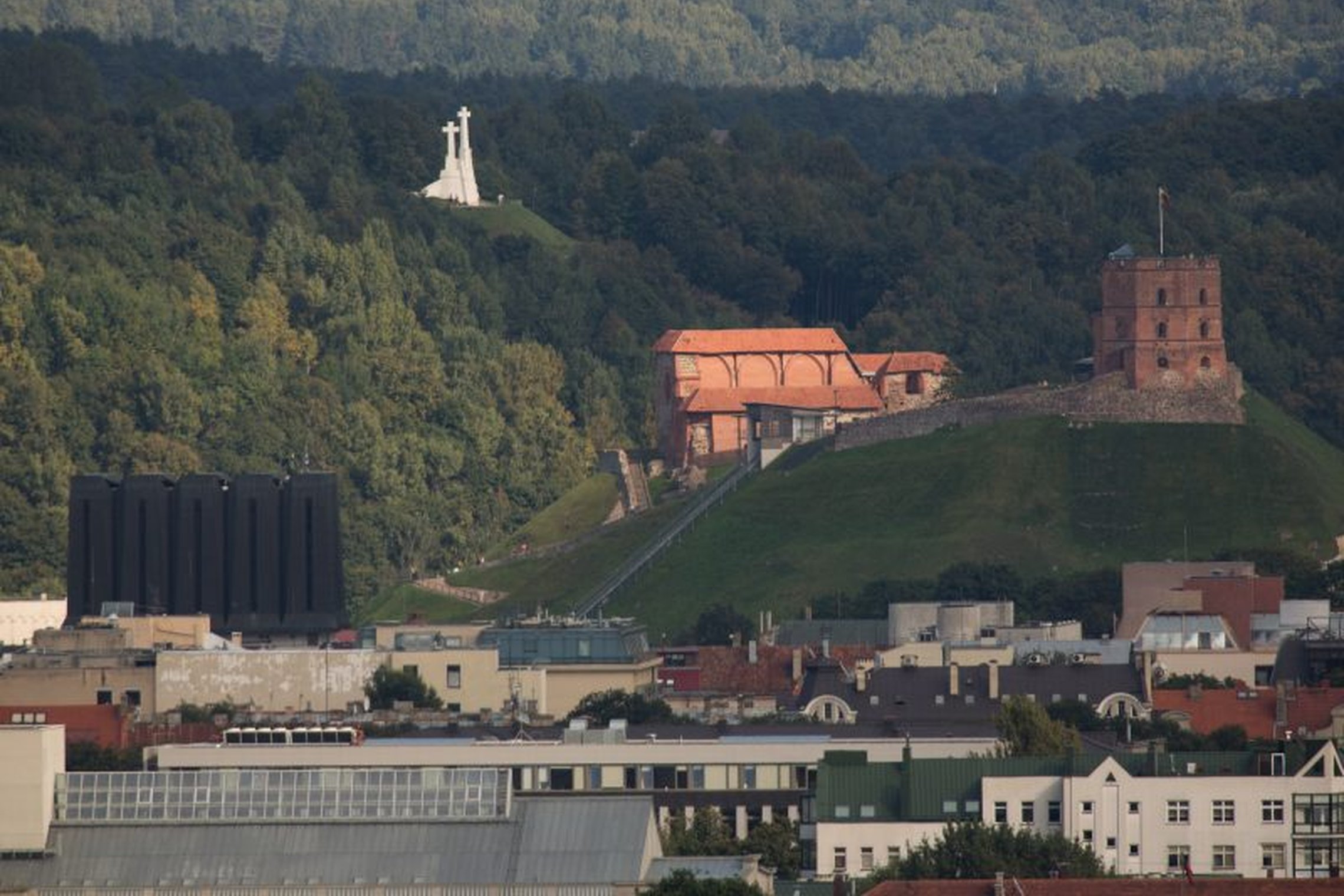 Vaizdas į Vilniaus pilių teritoriją. Matosi Vilniaus pilių kalnai, kurie apžėlę medžiais ir krūmais. Gražiai baltuoja Trijų Kryžių paminklas. Gedimino pilies griuvėsiais kaip ant delno. Kalnų papėdėje, nuotraukos priekyje - miesto namų stogai.