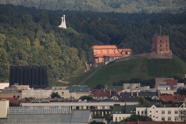 Vaizdas į Vilniaus pilių teritoriją. Matosi Vilniaus pilių kalnai, kurie apžėlę medžiais ir krūmais. Gražiai baltuoja Trijų Kryžių paminklas. Gedimino pilies griuvėsiais kaip ant delno. Kalnų papėdėje, nuotraukos priekyje - miesto namų stogai.