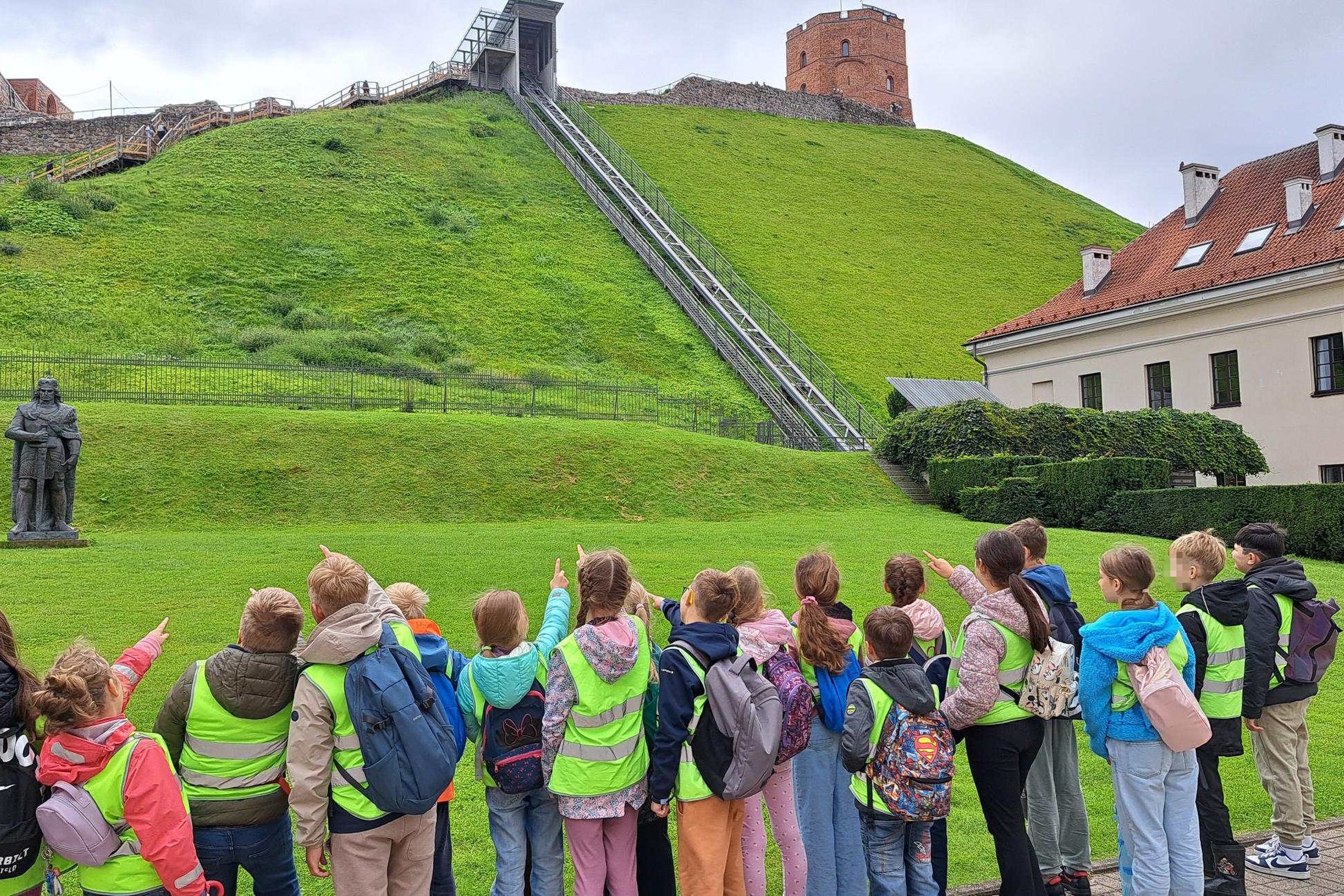 Vaikai su ryškiaspalvėmis liemenėmis stovi Vilniaus pilių teritorijoje, rodo į Gedimino pilies bokštą ant kalno, šalia matyti Gedimino skulptūra ir funikulierius.