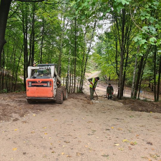 Du vyrai su kastuvais tvarko taką. Šalia stovi nedideis ekskavatoriukas. Už darbininkų matosi vielinė tvora - remonto darbai yra aptverti. Ankstyvas ruduo, medžiai dar labai žali.