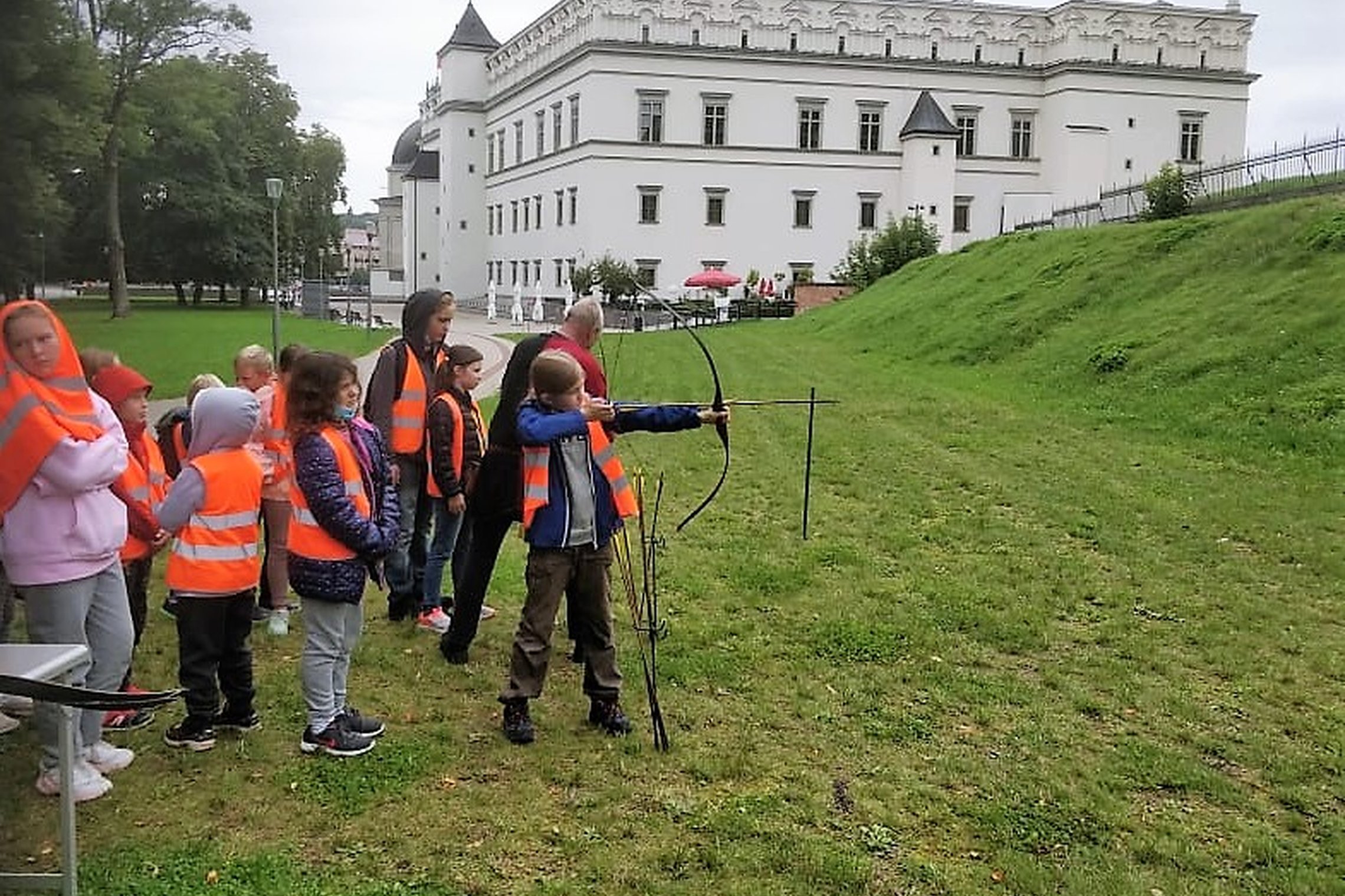 Būrelis vaikų šaudo iš lankų Gedimino kalno papėdėje. Kairėje matosi Valovų rūmai. Vaikai dėvi ryškias ir vienodas padidinto matomumo liemenes.