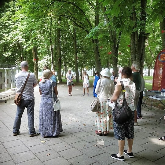 Pilių parke vyksta Vilniaus pilių paukščių nuotraukų parodos atidarymas. Susirinkę būrelis žmonių. Yra išdėliotos kėdės ir stalas. Aplinkui seni medžiai.