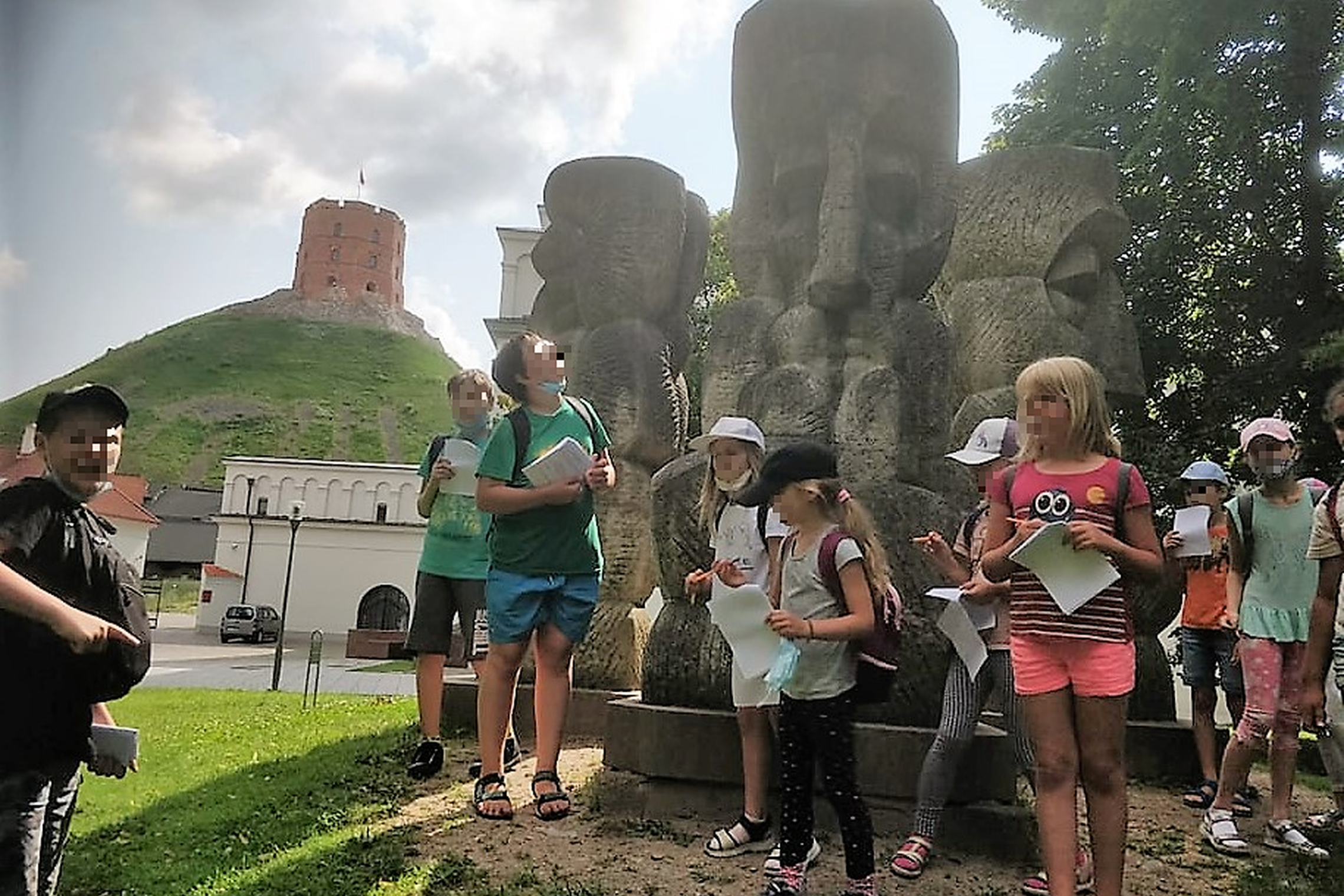 Šalia skulptūros TRYS KARALIAI stovi stovyklautojai. Jie sprendžia orientacinio žaidimo užduotis, Nuotraukos fone matosi Gedimino pilies kalnas ir bokštas.