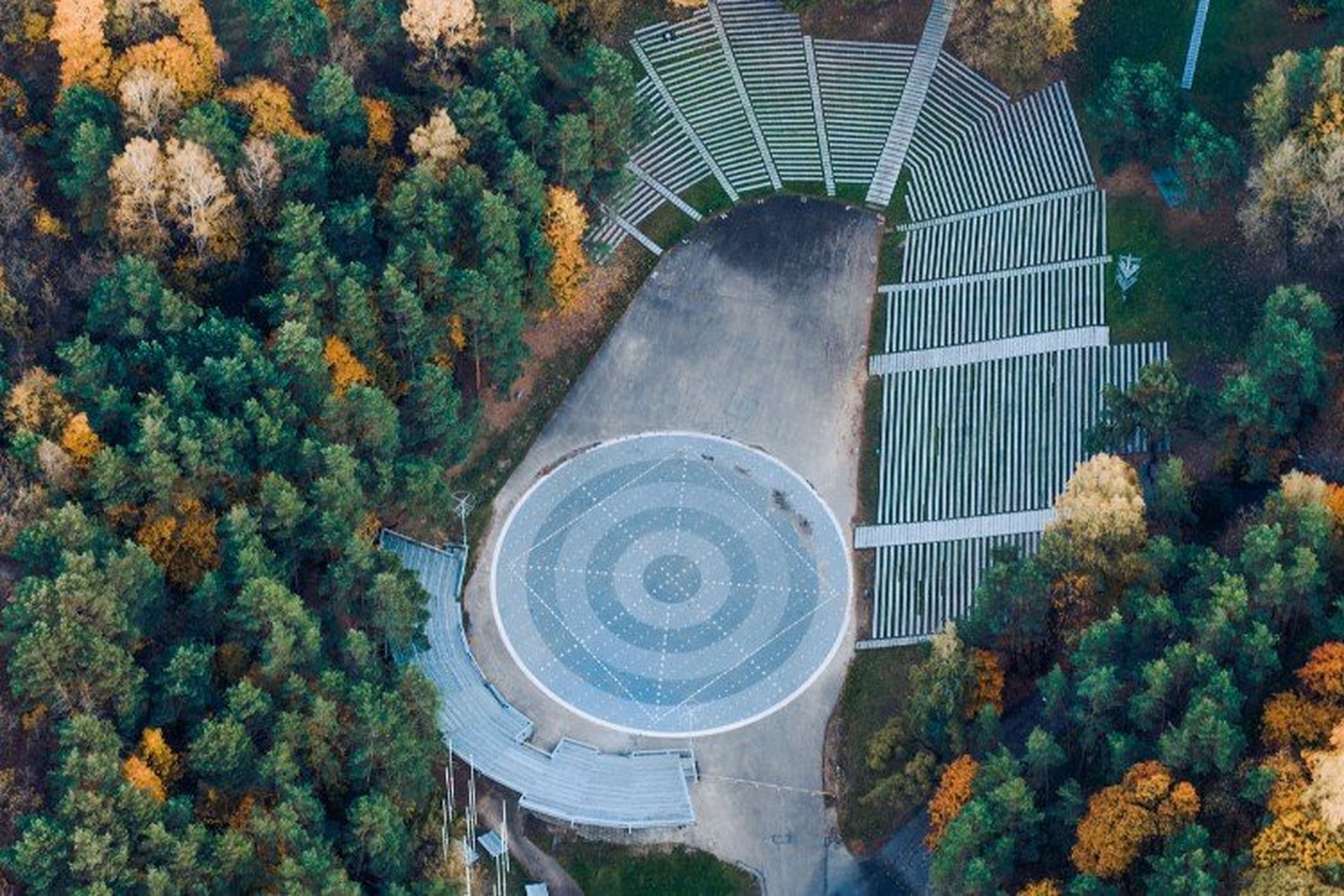 Dainų slėnis su medine estrada ir apvalia šokių aiškštele nufotografuotas iš drono - vaizdas iš viršaus. Aplinkui Dainų slėnį - rudenėjantys medžiai, nusidažę geltonais atspalviais. Ankstyvas ruduo.
