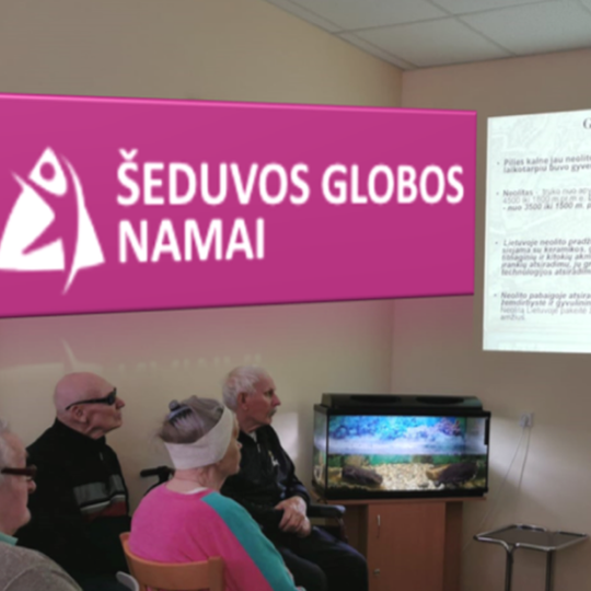 Nuotraukoje žmonės kurie stebi skaidres, rodomas televizoriaus ekrane. Žmonės susikaupę, keli yra su akiniais. Nuotraukos centre yra žuvyčių akvariumas. Tai senjorų globos namai.