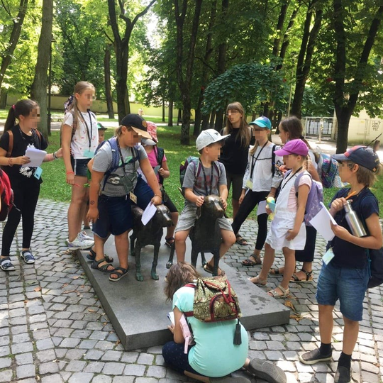 Būrys vaikų sėdi ant lietuvių skalikų skulptūros. Skulprūra yra Pilių parke.