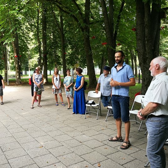 Pilių parke vyksta Vilniaus pilių paukščių nuotraukų parodos atidarymas. Susirinkę būrelis žmonių. Yra išdėliotos kėdės ir stalas. Aplinkui seni medžiai.