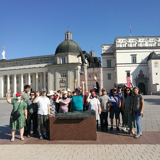 Didelė ekskursijos grupė stov aplinkui bronzinį maketą. Už jų Vilniaus katedra ir Valdovų rūmai.