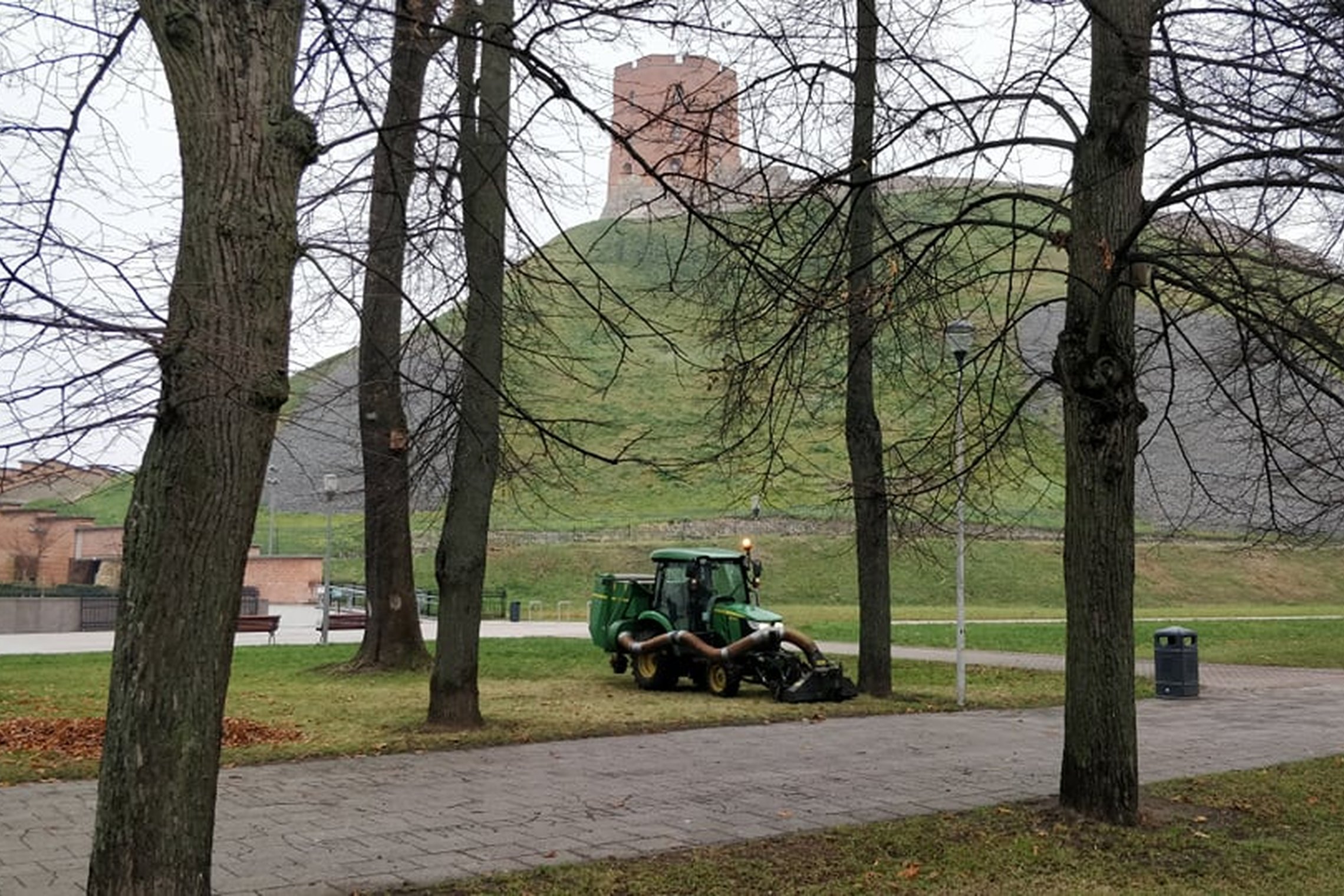 Senas parkas, kurio vejomis važiuoja nedidelis traktorius. Jis su specialiu įrenginiu siurbia rudeninius lapus. Tai paskutiniai lapai, nes ant medžių nėra nei vieno lapo. Nuotraukos antrame plane matosi Gedimino kalnas ir Gedimino pilies bokštas.