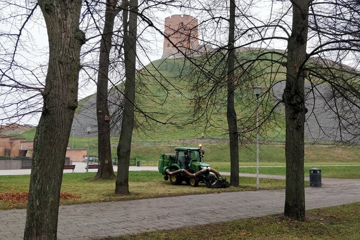 Senas parkas, kurio vejomis važiuoja nedidelis traktorius. Jis su specialiu įrenginiu siurbia rudeninius lapus. Tai paskutiniai lapai, nes ant medžių nėra nei vieno lapo. Nuotraukos antrame plane matosi Gedimino kalnas ir Gedimino pilies bokštas.