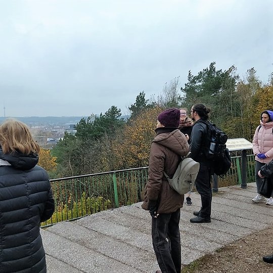 Žmonės stovi Trijų Kryžių papėdėje ir žiūri į Vilnių. Už jų yra med-iai, kurie pasipuošę rudeninėmis spalvomis.