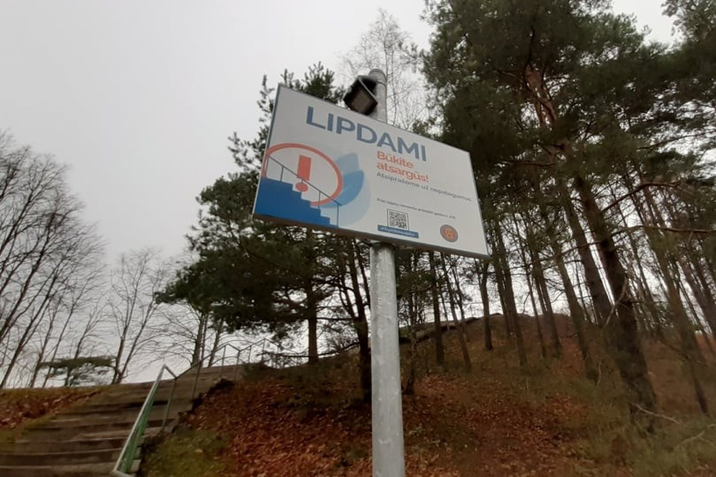 Kelių pušų fone yra iškelta ant ilgo koto lentelė - LIPDAMI BŪKITE ATSARGŪS. Tai perspėjimas žmonėms, nes Trijų Kryžių kalno laiptai laukia tvarkymo darbų.